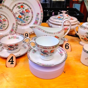 Könnte beinhalten: Ein weißes Porzellan-Teeservice mit Blumen- und Vogelmuster. Das Set umfasst Tassen, Untertassen, Teller, eine Sauciere und eine Servierschüssel mit Deckel. Die Ränder sind mit einem braunen Rand und floralen Akzenten versehen.