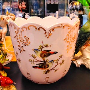 May include: White ceramic planter with a scalloped rim and gold accents. The planter features a detailed illustration of two colorful birds perched on a branch. A decorative piece for indoor plants.