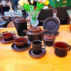 May include: A brown ceramic tea set, including cups, saucers, teapots, and a creamer, arranged on a wooden table. A sugar bowl and a decanter are also present. A vase of tulips adds a splash of colour.