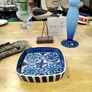 以下が含まれることがあります： 青い花柄と白黒のストライプの縁取りが施された正方形のセラミック皿。赤い頭と黒い翼を持つ木製の鳥の彫刻が近くに立っています。背の高い青いガラスの花瓶も存在します。