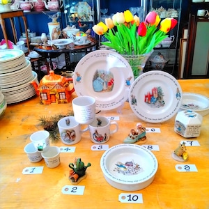 Puede incluir: Una colección de tazas de té, platos y figuritas con temática de Peter Rabbit. Los artículos están dispuestos sobre una mesa de madera con etiquetas numeradas. Los artículos son todos blancos con detalles azules y rojos. También hay algunas tulipanes en un jarrón en el fondo.