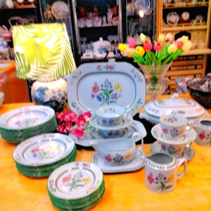 May include: A set of floral-patterned porcelain tableware, including plates, cups, saucers, a serving platter, and a gravy boat. The plates have green rims, and the floral designs feature pink, yellow, and blue flowers. A lamp with a green shade is in the background.