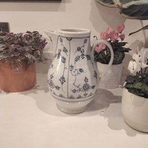 Może przedstawiać: Biały dzbanek ceramiczny z niebiesko-białym wzorem kwiatowym. Dzbanek ma szeroką podstawę i wąski dzióbek. Ma uchwyt po boku.