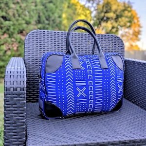 Può includere: Una borsa tote blu e bianca a fantasia con dettagli e manici in pelle nera. La borsa rettangolare è appoggiata su una sedia di vimini nera, con uno sfondo verde sfocato.