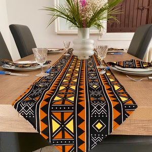 Peut inclure: Un chemin de table noir et orange avec un motif géométrique. Le chemin de table est posé sur une table en bois avec deux chaises et un vase de fleurs au centre.
