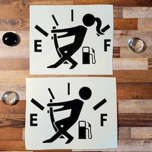 May include: Two black and white stickers depicting a fuel gauge with a person pushing the needle towards the full mark. The top sticker shows a woman with a ponytail, while the bottom sticker shows a man. Both stickers have the letters "E" and "F" on either side of the gauge.