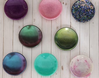 May include: A collection of seven small, round dishes in various colors and finishes. The colors include purple, pink, green, blue, and a glittery navy. The dishes are arranged against a light-colored, wood-grain background.
