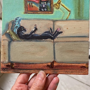 Könnte beinhalten: Ein quadratisches Leinwandgemälde zeigt eine schwarze Katze, die auf einem beigen Sofa liegt und ein kleines rechteckiges Gerät hält. Ein gerahmtes Bild einer schwarzen Katze hängt an der hellgrünen Wand über dem Sofa, neben einer gelben Lampe.