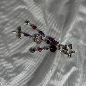 May include: A handmade beaded charm featuring a dragonfly, a flower, a spiral, and star-shaped charms. The charm has purple and pink beads. The charm is on a white, textured background.
