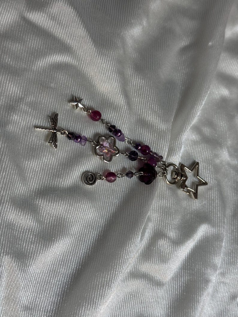 May include: A handmade beaded charm featuring a dragonfly, a flower, a spiral, and star-shaped charms. The charm has purple and pink beads. The charm is on a white, textured background.