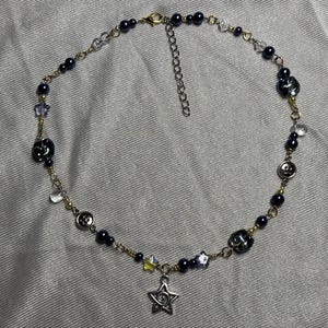 May include: A beaded necklace featuring dark navy blue spherical beads, clear star-shaped beads, and silver-toned metal buttons strung on a gold-toned chain. A silver-toned star charm hangs from the center, and a metal lobster clasp with an extender chain.