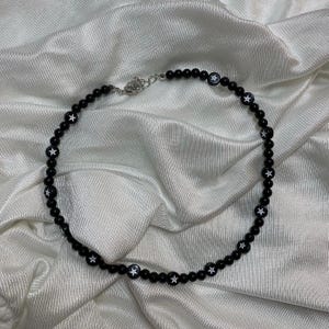 Könnte beinhalten: Eine schwarze Perlenkette mit weißen Sternanhängern. Die Kette liegt auf einem weißen Stoffhintergrund.