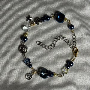 K&ouml;nnte beinhalten: Ein Bettelarmband mit dunkelblauen Perlen, kleinen cremefarbenen Perlen und silberfarbenen Metallakzenten. Das Armband enth&auml;lt Anh&auml;nger in Form eines Sterns, einer Spirale und eines Knopfes, die durch eine silberfarbene Kette mit Karabinerverschluss verbunden sind.