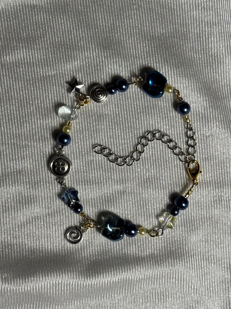K&ouml;nnte beinhalten: Ein Bettelarmband mit dunkelblauen Perlen, kleinen cremefarbenen Perlen und silberfarbenen Metallakzenten. Das Armband enth&auml;lt Anh&auml;nger in Form eines Sterns, einer Spirale und eines Knopfes, die durch eine silberfarbene Kette mit Karabinerverschluss verbunden sind.