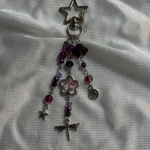 May include: A silver-toned keychain with a star-shaped clasp. Dangling from the clasp are strands of purple and clear beads, a flower charm, a dragonfly charm, a star charm, and a spiral charm. The beads are various sizes and shades of purple.