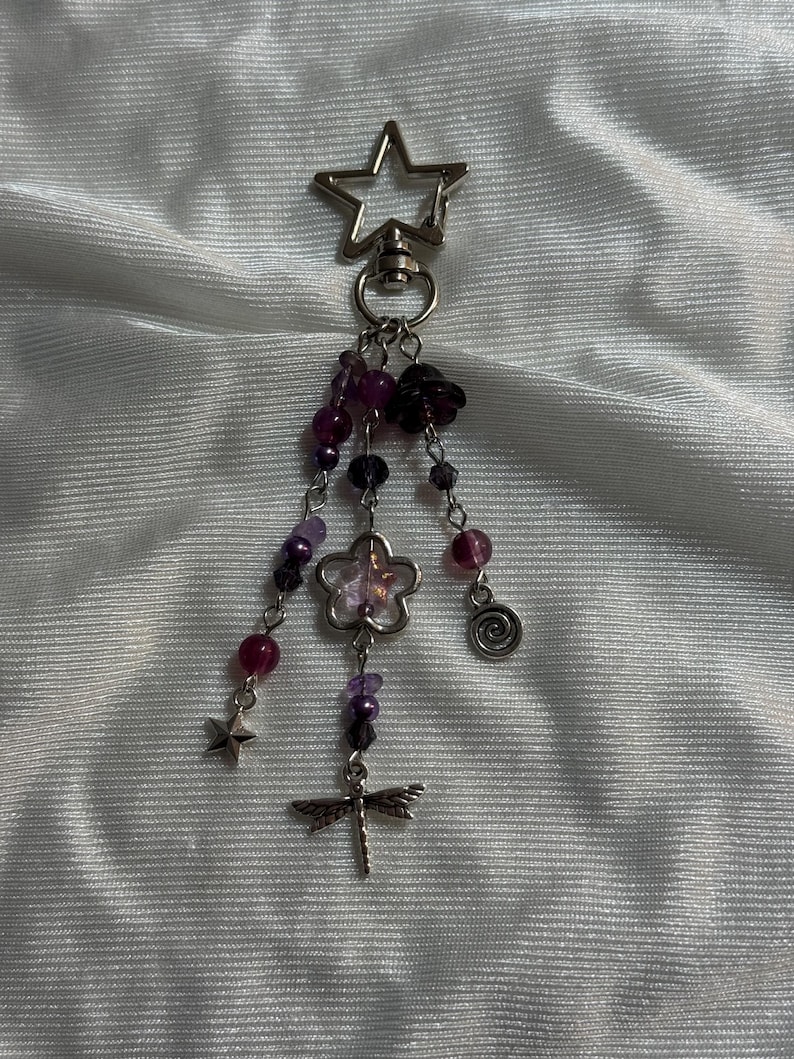May include: A silver-toned keychain with a star-shaped clasp. Dangling from the clasp are strands of purple and clear beads, a flower charm, a dragonfly charm, a star charm, and a spiral charm. The beads are various sizes and shades of purple.