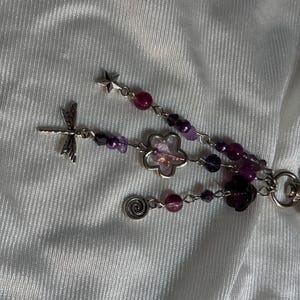 May include: A beaded keychain with a dragonfly charm, star charm, and flower charms. The keychain features purple and pink beads, with a silver-colored metal chain and charms. The background is a white, textured fabric.