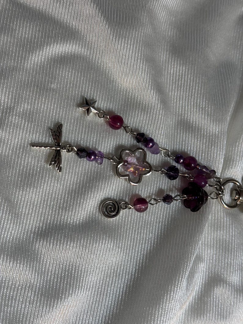 May include: A beaded keychain with a dragonfly charm, star charm, and flower charms. The keychain features purple and pink beads, with a silver-colored metal chain and charms. The background is a white, textured fabric.