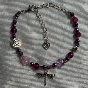 May include: A beaded bracelet featuring purple and pink beads, with a silver dragonfly charm. The bracelet includes a spiral bead, star-shaped beads, and a heart-shaped charm on the adjustable chain.
