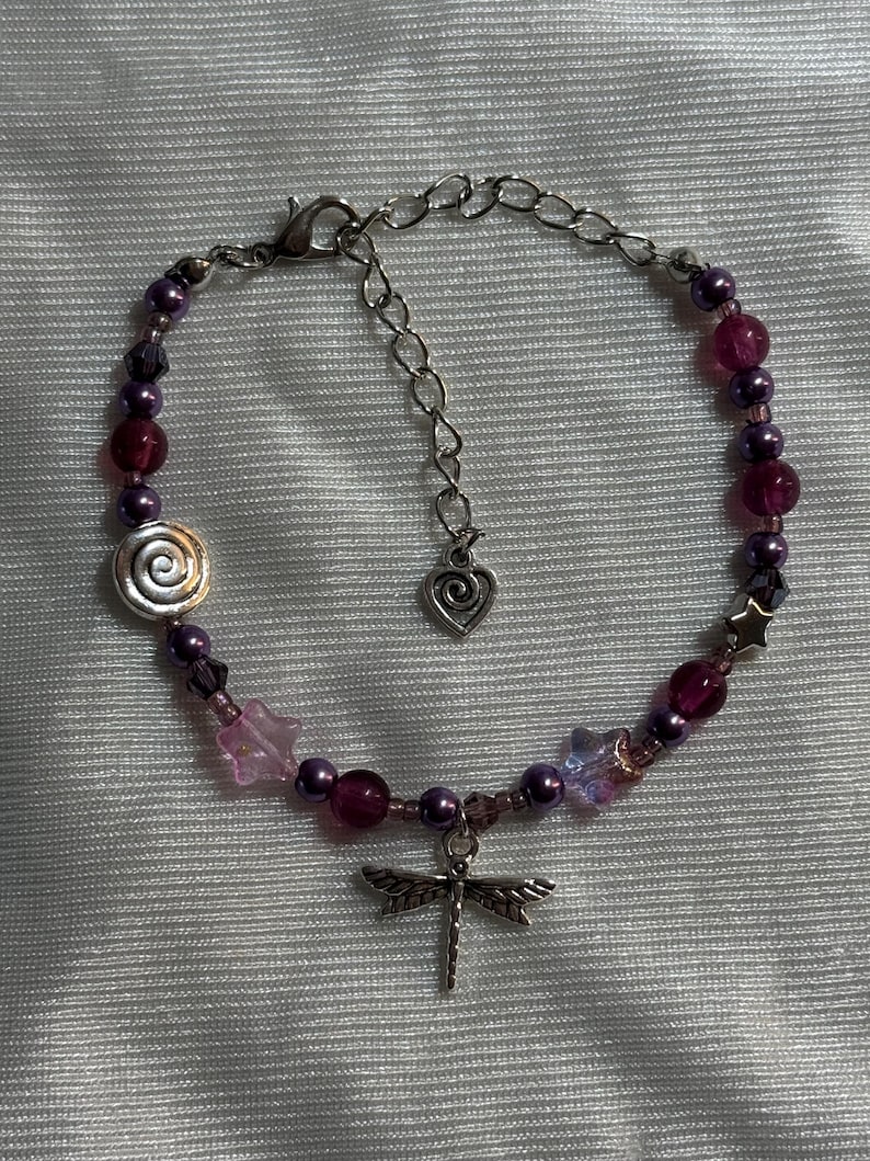 May include: A beaded bracelet featuring purple and pink beads, with a silver dragonfly charm. The bracelet includes a spiral bead, star-shaped beads, and a heart-shaped charm on the adjustable chain.