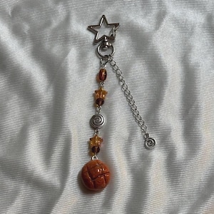 Könnte beinhalten: Ein dekorativer Schlüsselanhänger mit sternförmigem Oberteil und Kette. Die Kette ist mit orangefarbenen Perlen, sternförmigen Charms und einem Spiral-Charm verziert. Am unteren Ende hängt ein runder, strukturierter, orangefarbener Charm.
