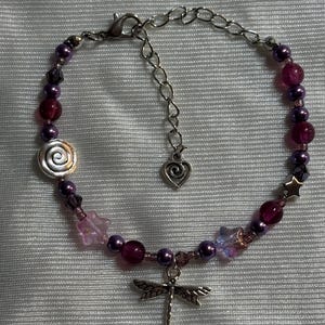 May include: A beaded bracelet featuring a dragonfly charm, spiral, and star-shaped accents. The bracelet incorporates various shades of purple beads and crystals, with a silver-toned chain and clasp. The bracelet is designed for a delicate and feminine aesthetic.
