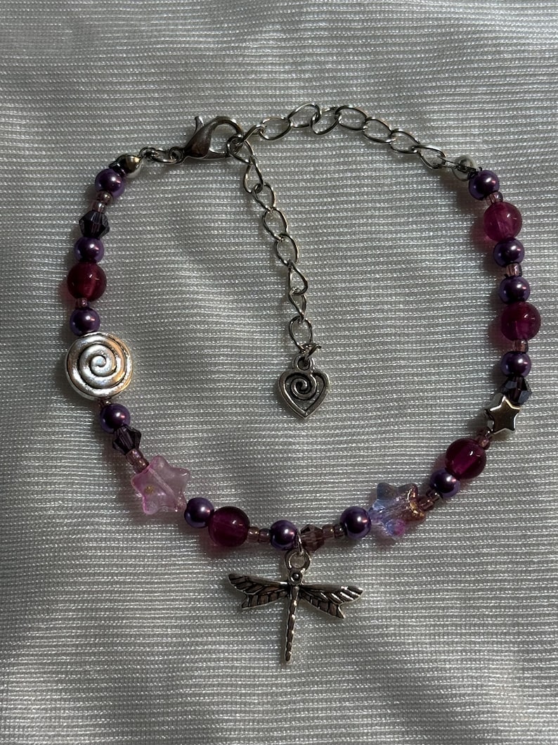 May include: A beaded bracelet featuring a dragonfly charm, spiral, and star-shaped accents. The bracelet incorporates various shades of purple beads and crystals, with a silver-toned chain and clasp. The bracelet is designed for a delicate and feminine aesthetic.