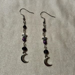 Könnte beinhalten: Ein Paar Ohrhänger mit silbernem Halbmond-Anhänger, Stern-Anhänger und dunkelvioletten Perlen. Die Ohrringe haben ein silbernes Haken- und Ketten-Design. Die Ohrringe werden auf einem hellen Stoff präsentiert.