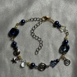 K&ouml;nnte beinhalten: Ein Armband mit dunkelblauen Perlen, durchscheinenden blauen Glasperlen, silberfarbenen Stern- und Spiralanh&auml;ngern sowie einem zentralen Vierlochknopf. Das Schmuckst&uuml;ck ist mit goldfarbenem Draht gefertigt und besitzt eine silberfarbene Kettenverl&auml;ngerung.