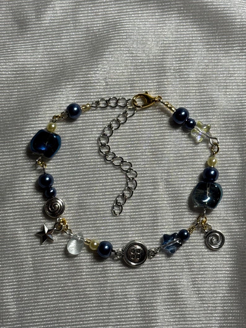 K&ouml;nnte beinhalten: Ein Armband mit dunkelblauen Perlen, durchscheinenden blauen Glasperlen, silberfarbenen Stern- und Spiralanh&auml;ngern sowie einem zentralen Vierlochknopf. Das Schmuckst&uuml;ck ist mit goldfarbenem Draht gefertigt und besitzt eine silberfarbene Kettenverl&auml;ngerung.