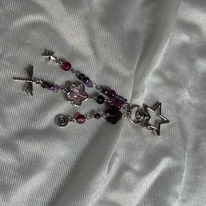 May include: A handmade keychain featuring a dragonfly, flower, star, and spiral charm. The keychain has purple and pink beads, with a silver-colored metal finish. The design is delicate and whimsical.