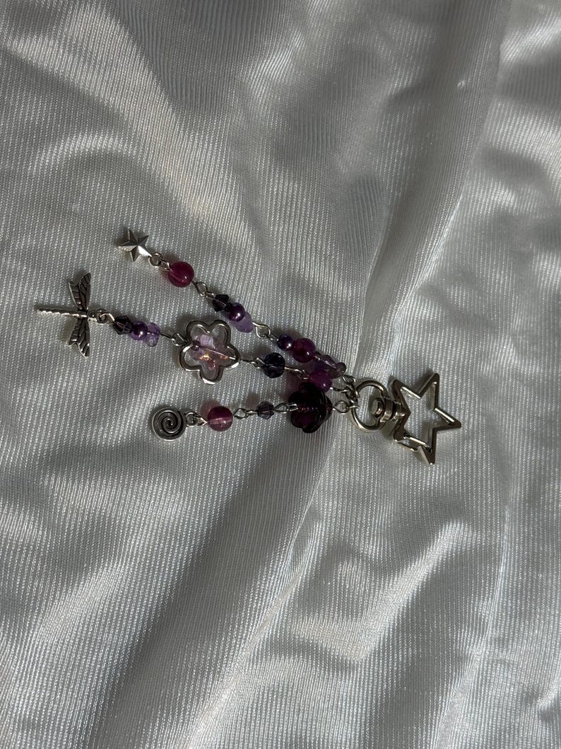 May include: A handmade keychain featuring a dragonfly, flower, star, and spiral charm. The keychain has purple and pink beads, with a silver-colored metal finish. The design is delicate and whimsical.