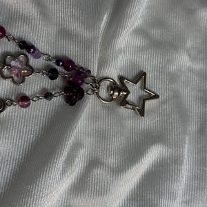 May include: A beaded keychain with purple and clear beads, silver-colored chain, and a star-shaped clasp. The keychain features flower-shaped charms and various shades of purple beads.