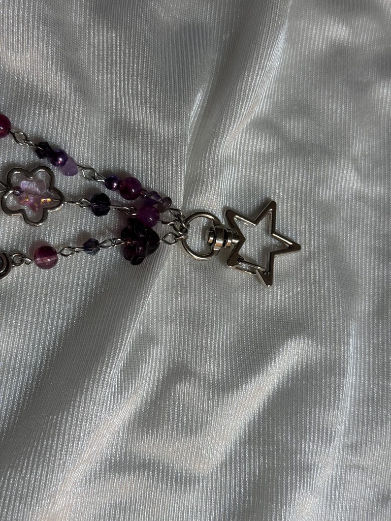 May include: A beaded keychain with purple and clear beads, silver-colored chain, and a star-shaped clasp. The keychain features flower-shaped charms and various shades of purple beads.