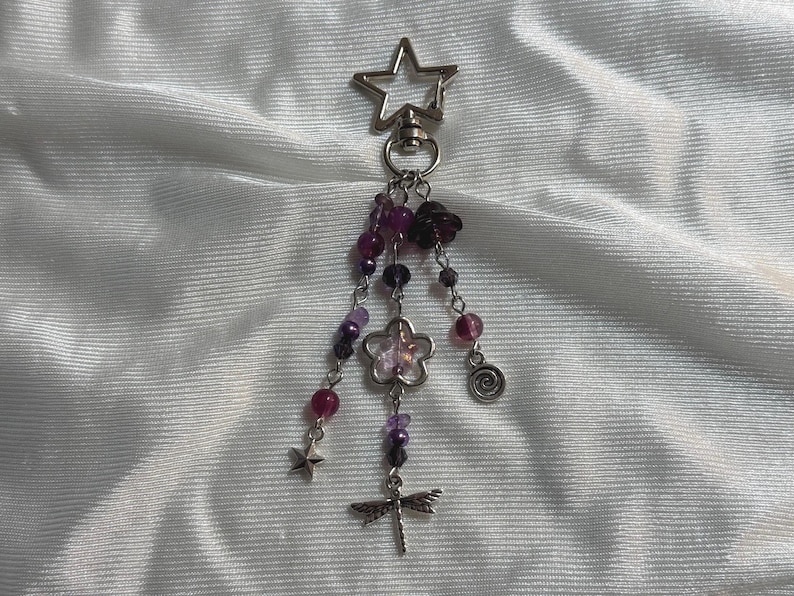 May include: A silver-toned keychain with a star-shaped clasp. Dangling from the clasp are strands of purple and clear beads, a flower charm, a dragonfly charm, a star charm, and a spiral charm. The background is a white, textured fabric.