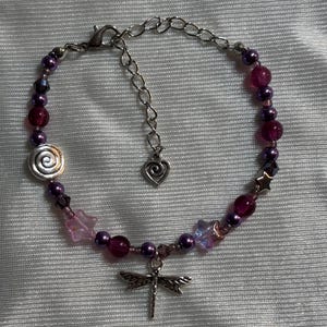 May include: A beaded bracelet featuring a dragonfly charm. The bracelet has a mix of purple and pink beads, with a silver spiral and star-shaped accents. The adjustable chain has a heart charm.