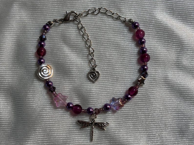 May include: A beaded bracelet featuring a dragonfly charm. The bracelet has a mix of purple and pink beads, with a silver spiral and star-shaped accents. The adjustable chain has a heart charm.