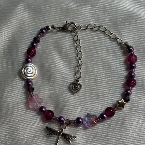 May include: A beaded bracelet with a silver-tone chain and clasp. The bracelet features a dragonfly charm, purple and pink beads, and star and flower-shaped accents. A spiral-shaped charm is also included.