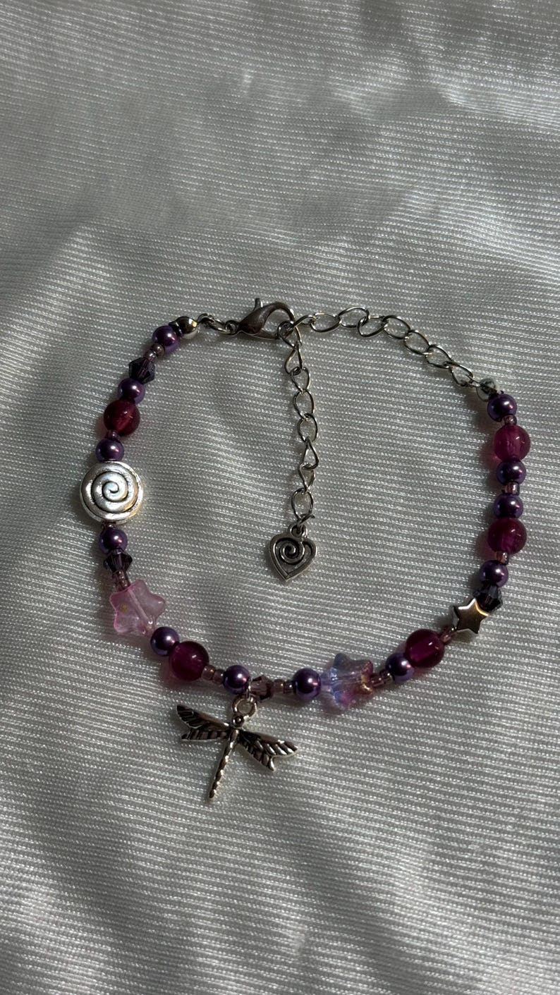May include: A beaded bracelet with a silver-tone chain and clasp. The bracelet features a dragonfly charm, purple and pink beads, and star and flower-shaped accents. A spiral-shaped charm is also included.