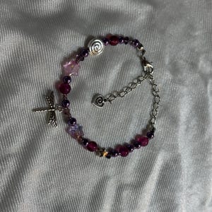 May include: A beaded bracelet with a dragonfly charm. The bracelet features a mix of purple and pink beads, with silver accents including a spiral, stars, and a heart-shaped clasp. The bracelet is on a white, textured surface.