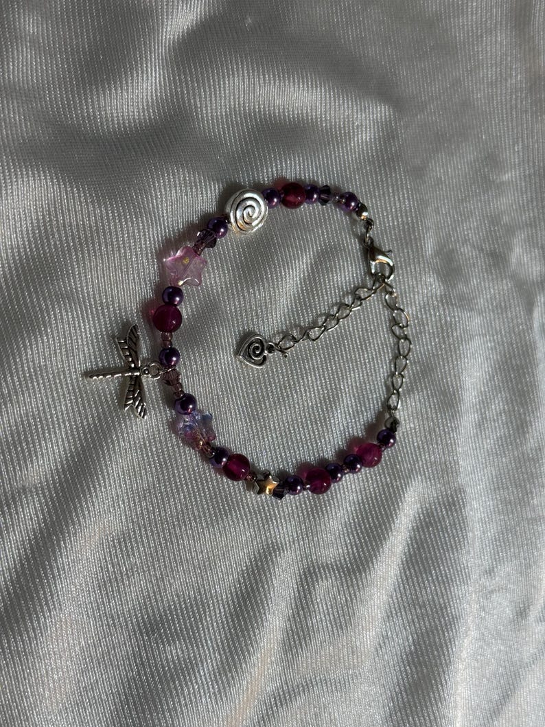 May include: A beaded bracelet with a dragonfly charm. The bracelet features a mix of purple and pink beads, with silver accents including a spiral, stars, and a heart-shaped clasp. The bracelet is on a white, textured surface.