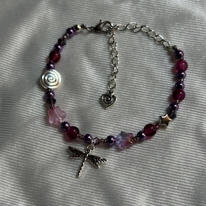 May include: A beaded bracelet featuring a dragonfly charm and various beads in shades of purple, pink, and silver. The bracelet includes a spiral bead, star-shaped beads, and a heart-shaped charm.
