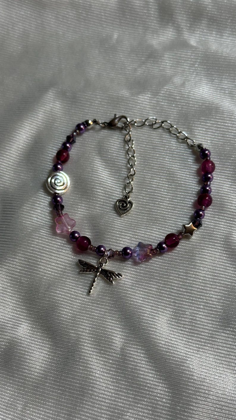 May include: A beaded bracelet featuring a dragonfly charm and various beads in shades of purple, pink, and silver. The bracelet includes a spiral bead, star-shaped beads, and a heart-shaped charm.