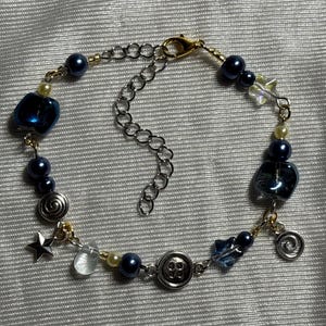 K&ouml;nnte beinhalten: Ein Perlenarmband mit dunkelblauen Perlen, klaren Glasperlen, silberfarbenen Sternanh&auml;ngern, spiralf&ouml;rmigen Metallanh&auml;ngern und einer knopff&ouml;rmigen Perle. Das Armband ist mit goldfarbenen Metallgliedern verbunden und hat einen silberfarbenen Karabiner.