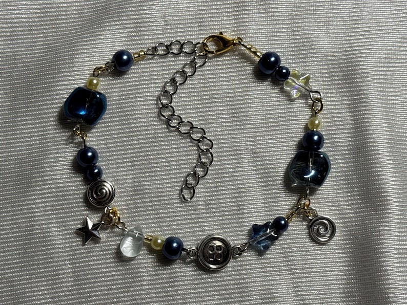 K&ouml;nnte beinhalten: Ein Perlenarmband mit dunkelblauen Perlen, klaren Glasperlen, silberfarbenen Sternanh&auml;ngern, spiralf&ouml;rmigen Metallanh&auml;ngern und einer knopff&ouml;rmigen Perle. Das Armband ist mit goldfarbenen Metallgliedern verbunden und hat einen silberfarbenen Karabiner.