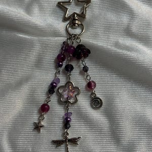 May include: A silver keychain with a star-shaped clasp. Dangling from the clasp are strands of purple beads, a flower charm, a dragonfly charm, a small star, and a spiral charm. The beads are various shades of purple.
