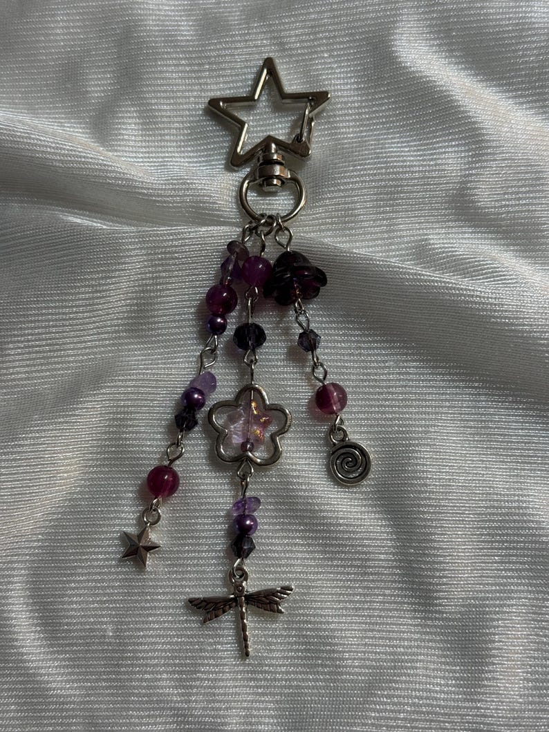 May include: A silver keychain with a star-shaped clasp. Dangling from the clasp are strands of purple beads, a flower charm, a dragonfly charm, a small star, and a spiral charm. The beads are various shades of purple.