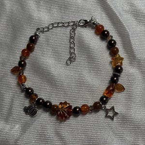 Op de afbeelding: Een kralenarmband met donkerbruine en doorschijnende barnsteenkleurige kralen, afgewisseld met zilverkleurige bedels in de vorm van een pompoen, een esdoornblad en een ster, afgewerkt met een zilverkleurige metalen ketting en karabijnsluiting.