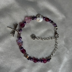 May include: A beaded bracelet featuring a dragonfly charm, star and flower accents, and a spiral bead. The bracelet incorporates purple and pink beads, with a silver-toned chain and clasp. The bracelet is designed for wrist wear.