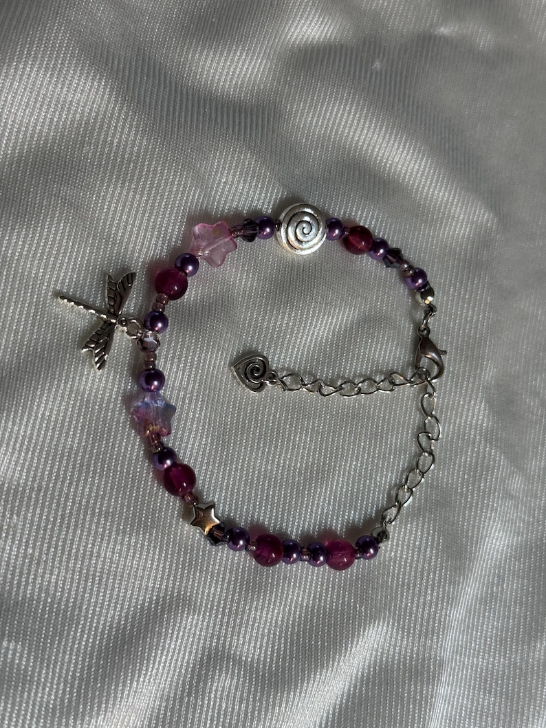 May include: A beaded bracelet featuring a dragonfly charm, star and flower accents, and a spiral bead. The bracelet incorporates purple and pink beads, with a silver-toned chain and clasp. The bracelet is designed for wrist wear.